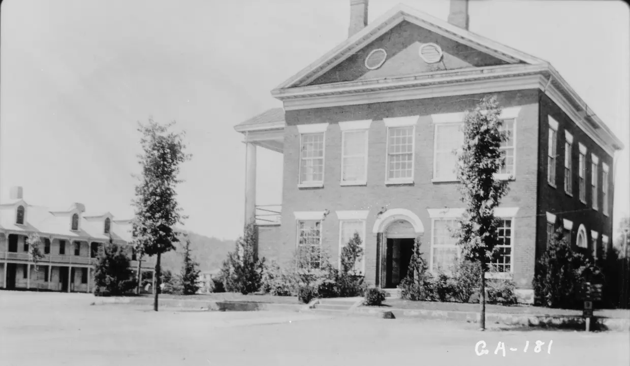 A 1936 black-and-white photograph of a two-storey brick building with a pedimented gable end, round and oval attic windows and two chimneys, standing on a bare unpaved square planted with young trees.