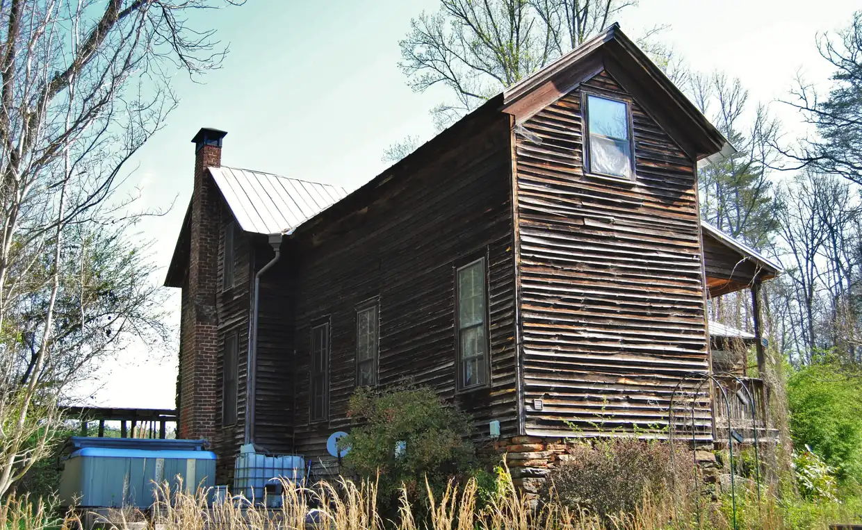 A tall weathered board-sided farmhouse with a metal roof and a small side porch, seen across a garden of tall grass and shrubs among bare spring trees.