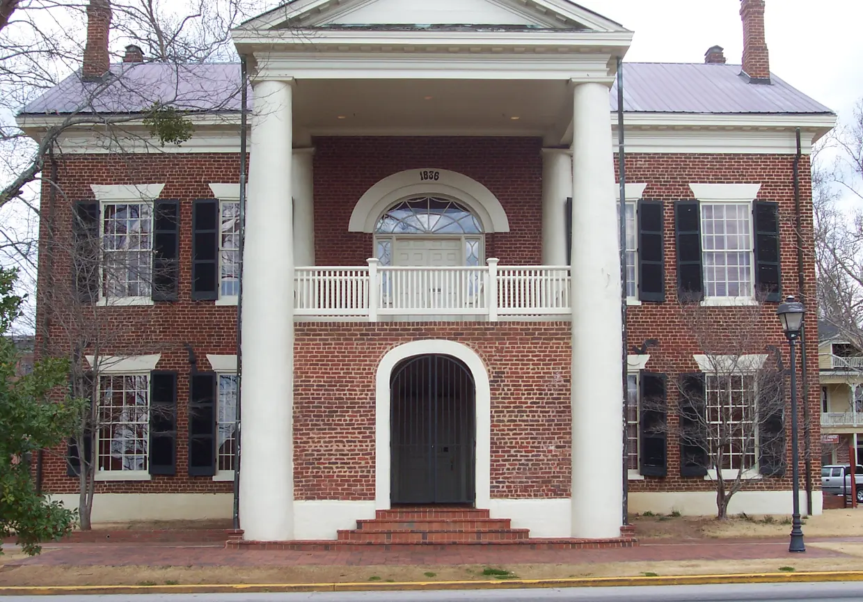 The red-brick front of the old courthouse with a tall white two-column portico, a balcony under a fanlight dated 1836, and an arched gated doorway at the top of brick steps.