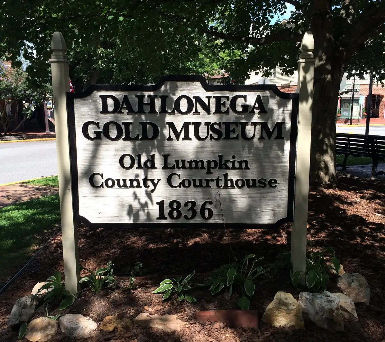 A white wooden sign reading Dahlonega Gold Museum, Old Lumpkin County Courthouse, 1836, set in a shaded garden bed with rocks and hostas.