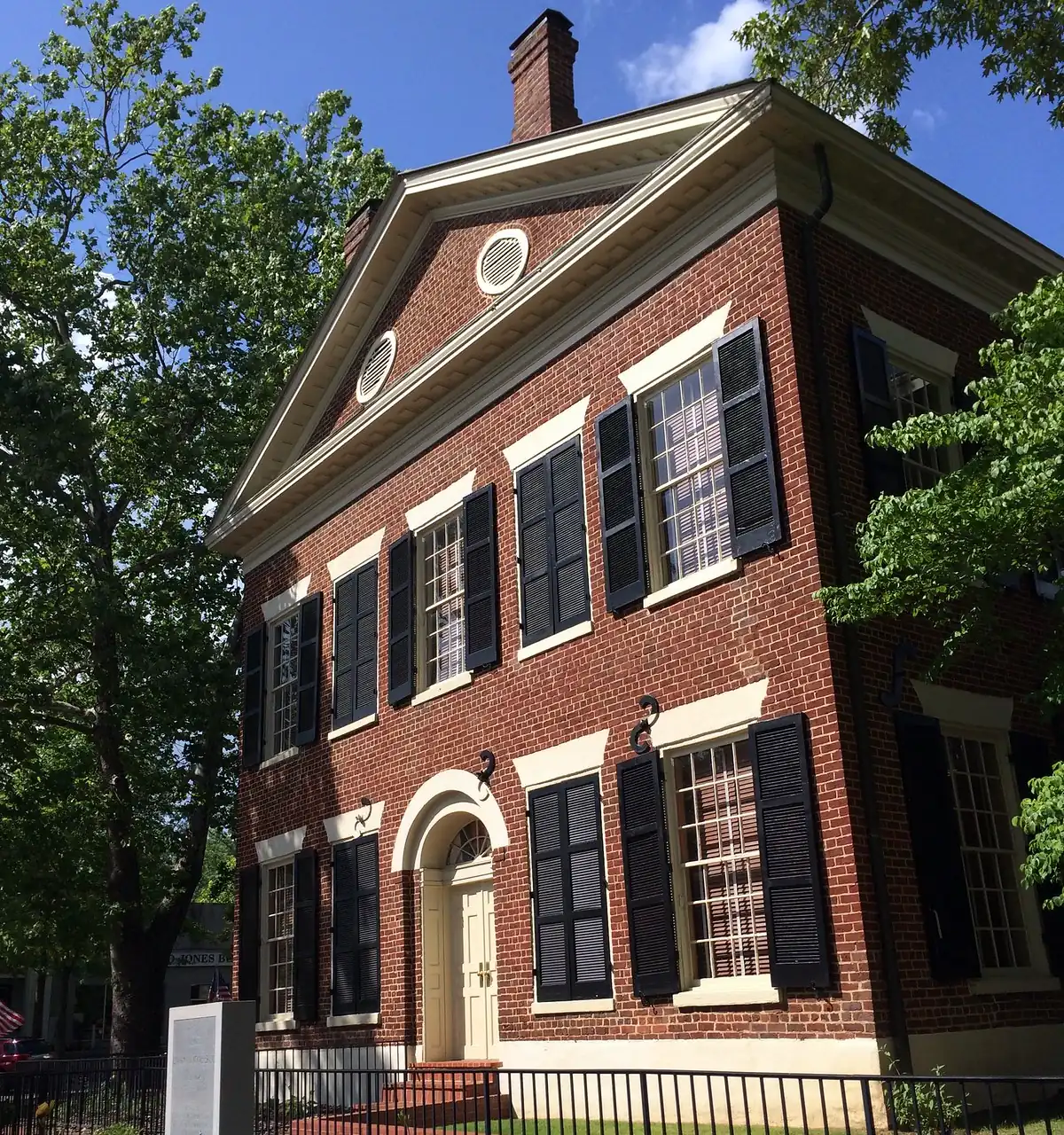 The pedimented gable end of a red-brick two-storey building with black shutters, an oval attic window and an arched doorway, behind an iron fence and garden under leafy summer trees.