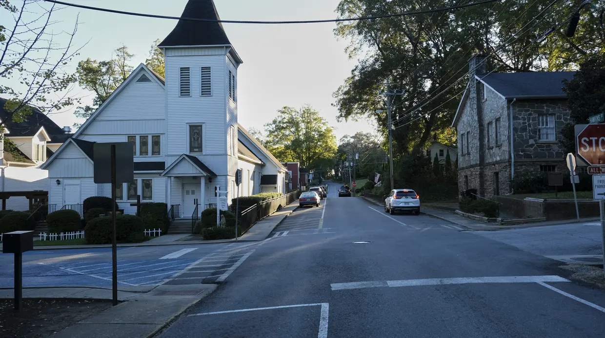 A quiet in-town street crossing: a white wooden church with a pointed steeple on the left, a grey stone building behind a stop sign on the right, and a tree-lined street with a few parked cars.