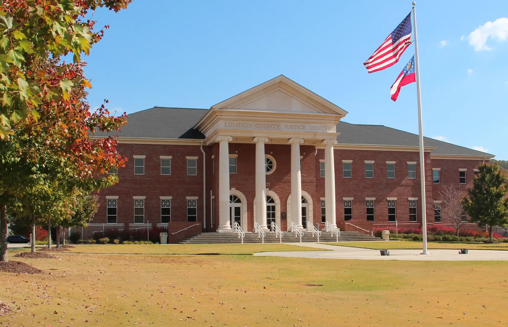 A long red-brick civic building with a white six-column portico and arched doorways, set back across a wide sunlit lawn, with the United States and Georgia flags on a tall pole and a tree in red autumn leaves at the left.