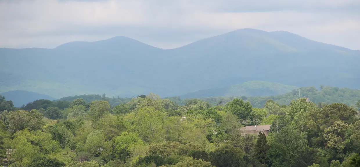 Two broad blue mountains on the horizon above a rolling green canopy of trees with a few rooftops showing, under a hazy sky.