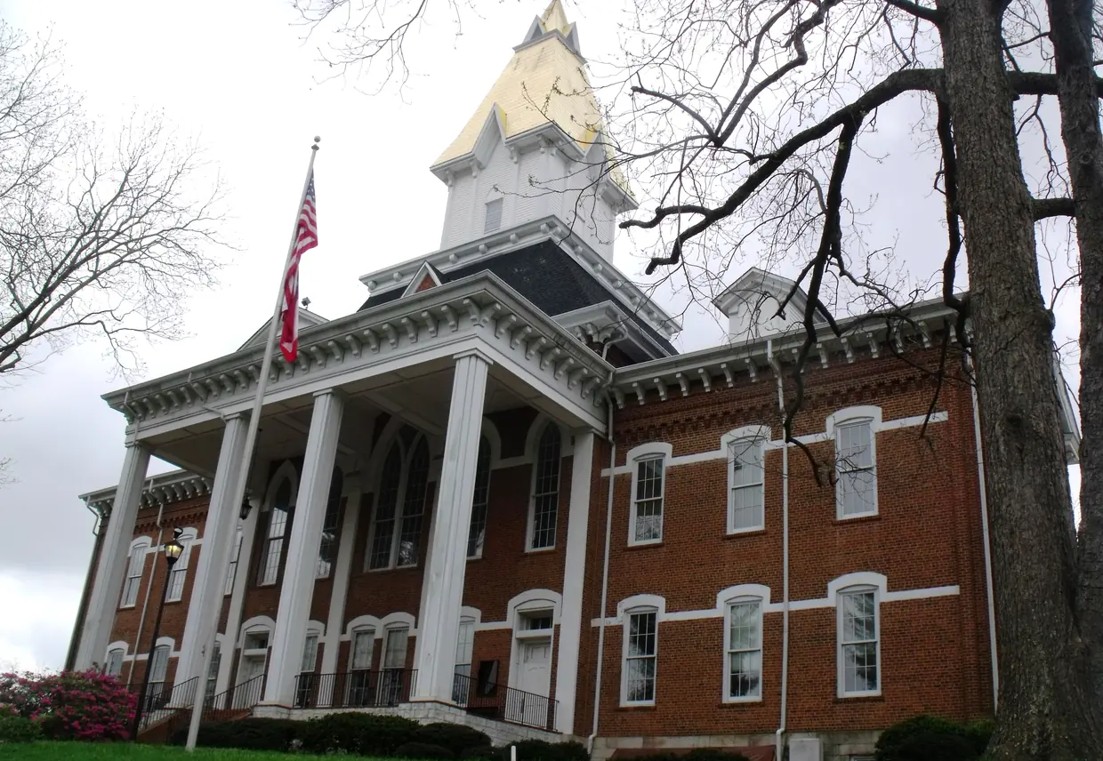A red-brick building with a tall portico of white columns and a white belfry topped by a gold-leafed steeple, seen from below with an American flag in front.