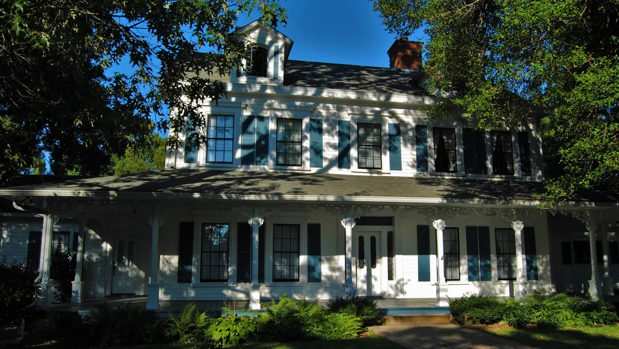 A large white two-storey house with a long columned wrap-around porch, black shutters and dormers, partly shaded by trees behind a lawn and shrubs.