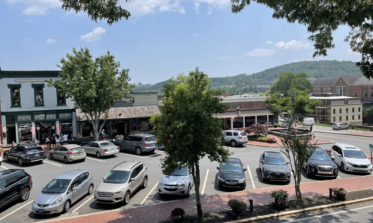 A high summer view over a parking lot full of cars to a row of storefronts on the square, leafy trees, and forested hills on the horizon under a blue sky with white clouds.