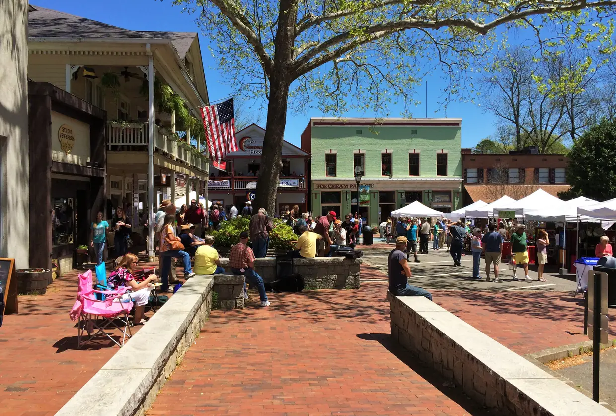 People strolling and sitting on low stone walls on a sunny brick plaza, with a flag-hung porch on the left, a green two-storey storefront and white vendor tents ahead, under a large spreading tree.