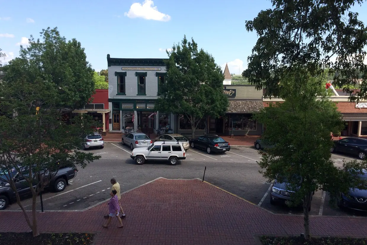 An elevated summer view over a brick crosswalk, where two people are walking, and a lot of parked cars to a row of two-storey storefronts among green trees.